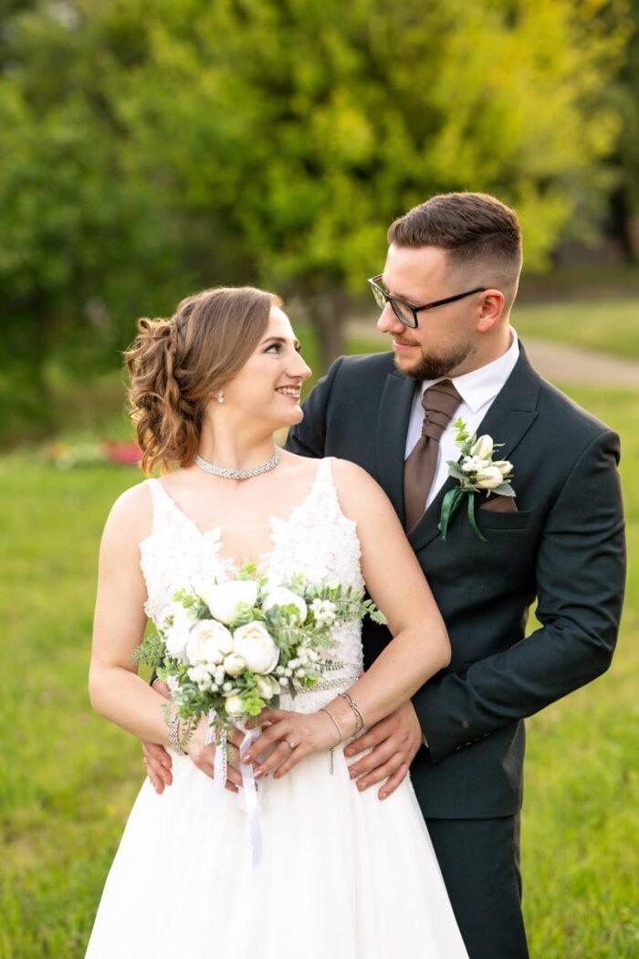 A boldog menyasszony és vőlegény egymásra mosolyog a zöld parkban. A menyasszony fehér csipkés ruhában és gyöngysoros nyaklánccal áll, kezében virágcsokrot tartva, míg a vőlegény sötét öltönyben hátulról öleli őt.