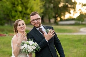 A friss házaspár mosolyogva mutatja gyűrűiket, miközben a háttérben a naplemente lágy fényei világítják meg a zöldellő parkot. Esküvő fotós Debrecenben