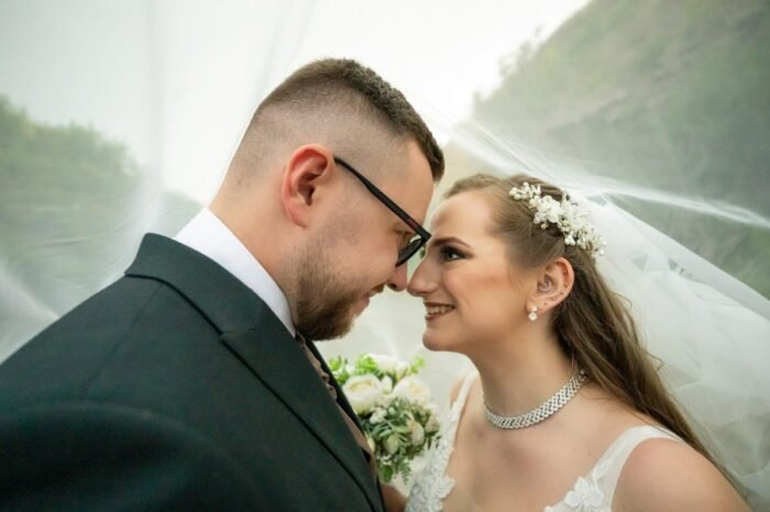 A boldog ifjú pár gyengéden egymásnak támasztja homlokát, miközben mosolyogva néznek egymás szemébe. A menyasszony finom gyöngyös hajdísszel és elegáns nyaklánccal ragyog, míg a vőlegény öltönyben sugározza a boldogságot. A fátyol lágyan keretezi a romantikus pillanatot, miközben a háttérben a természet halvány kontúrjai láthatók.