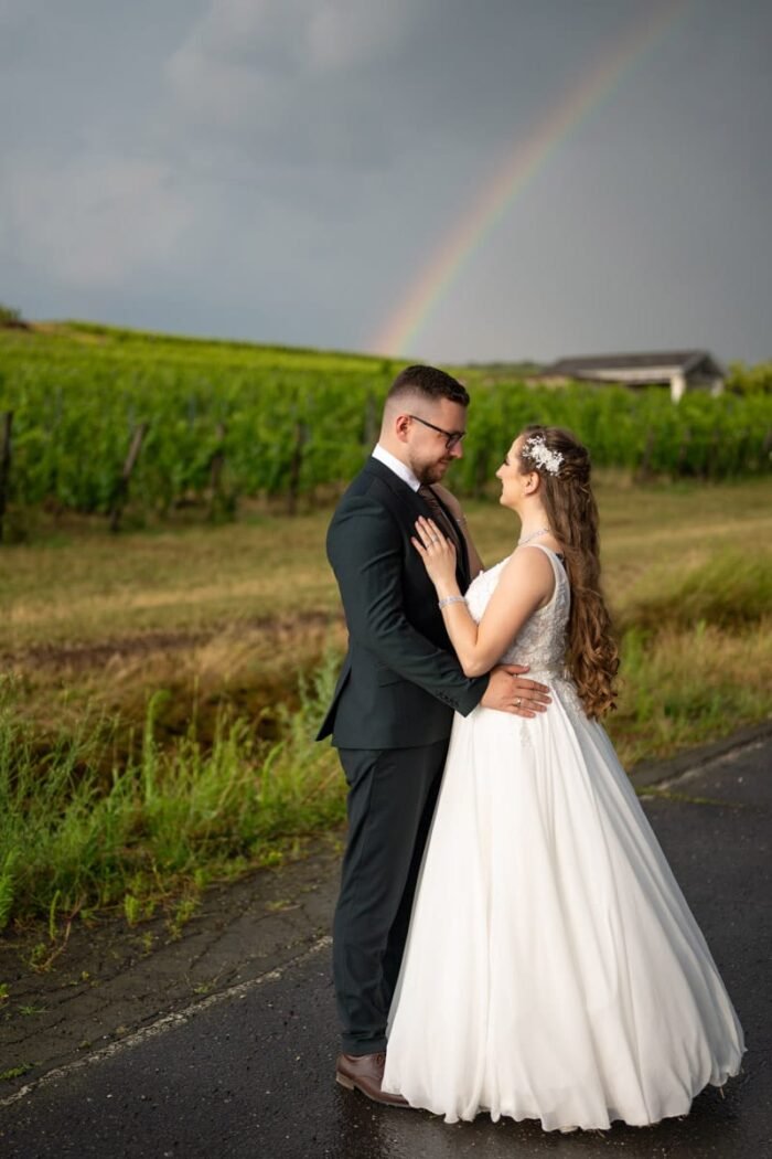 A menyasszony és a vőlegény gyengéden ölelik egymást egy eső utáni, szőlőskerti környezetben. Az égbolt drámai hátteret biztosít a szivárvánnyal, amely szimbolikusan az új kezdetet és a reményt jelképezi. A menyasszony finoman a vőlegény vállán nyugtatja kezét, miközben boldogan néznek egymásra. Az alkalom romantikus és idilli, a természet szépsége tökéletesen harmonizál az ifjú pár örömével.