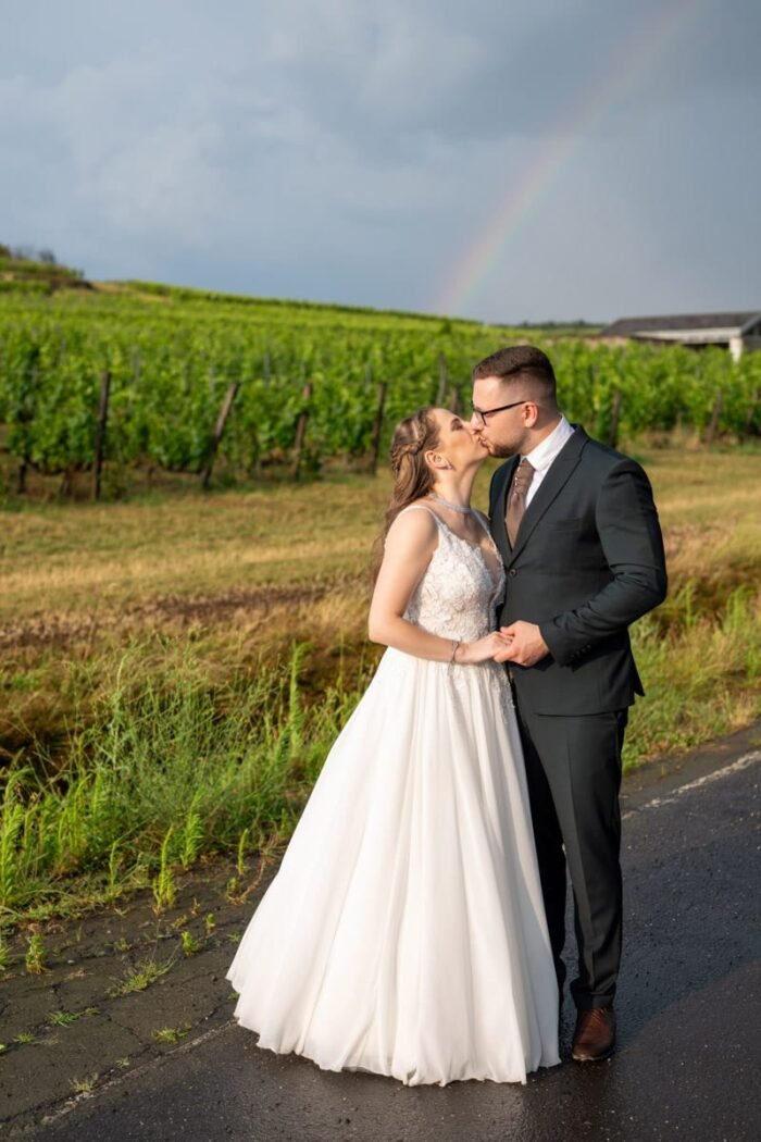 A menyasszony és a vőlegény romantikusan megcsókolják egymást egy eső utáni, szőlőskerti tájon. A háttérben egy lenyűgöző szivárvány ível az égbolton, amely még különlegesebbé teszi a pillanatot. A menyasszony kezét gyengéden a vőlegény kezében tartja, miközben esküvői ruhája elegánsan terül el körülötte. A természet nyugalma és szépsége tökéletes összhangban van a pár boldogságával.