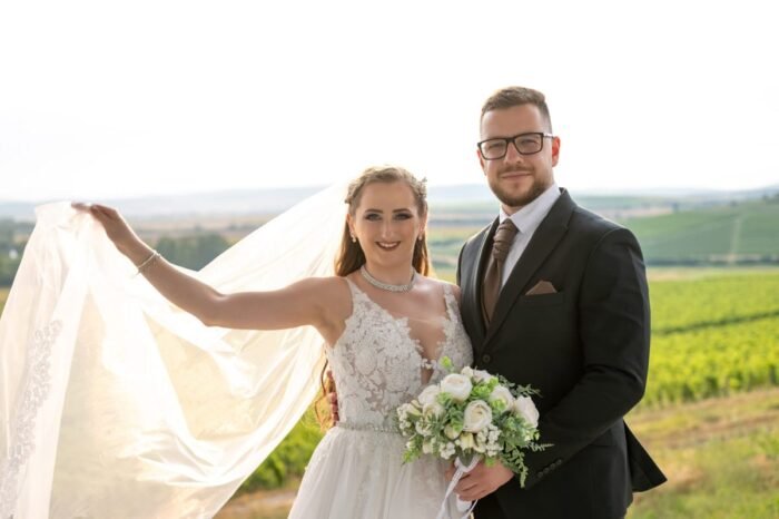 Egy boldog esküvői pár áll a szőlőskertek között, a naplemente lágy fényében. A menyasszony bájos mosollyal a fátylát emeli a szélben, míg a vőlegény mellette magabiztosan áll, öltönyben és nyakkendőben. A menyasszony kezében gyönyörű fehér rózsákból és zöld növényekből álló csokor található. A háttérben dombok és szőlőtőkék rajzolódnak ki, amelyek romantikus hangulatot kölcsönöznek a képnek.