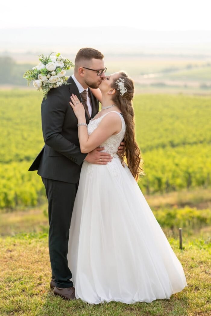 Egy boldog házaspár csókolózik a szőlőtőkék között. A vőlegény elegáns fekete öltönyben, kezében a menyasszony fehér rózsákból álló csokrával áll, miközben gyengéden öleli a menyasszonyt. A menyasszony csipkés fehér esküvői ruhát visel, haja hullámosan omlik a vállára. A háttérben a dombos táj és a zöldellő szőlőültetvények láthatók, amelyek idilli hangulatot teremtenek.