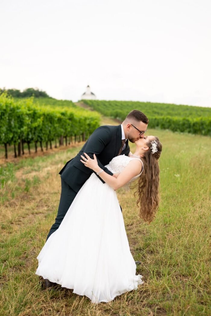 A vőlegény elegáns fekete öltönyben, menyasszonya gyönyörű fehér esküvői ruhában szenvedélyes csókot váltanak, miközben a vőlegény finoman hátradönti párját. A hátteret szőlőtőkék és egy távoli kápolna sziluettje díszíti, romantikus hangulatot kölcsönözve a jelenetnek.