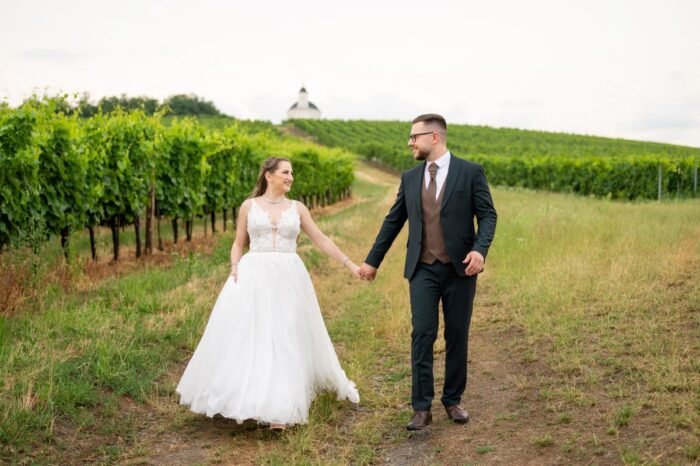 Egy mosolygó menyasszony és vőlegény kézen fogva sétálnak a szőlőtőkék között, romantikus környezetben. A menyasszony elegáns fehér ruhát, a vőlegény barna mellénnyel kiegészített fekete öltönyt visel. Esküvő videó Debrecen