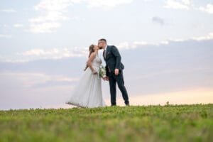 Egy menyasszony és vőlegény romantikusan megcsókolja egymást egy mezőn, a háttérben gyönyörű naplementés égbolttal. A menyasszony hófehér ruhát és csokrot tart, míg a vőlegény elegáns sötét öltönyben áll mellette. Az égbolt lágy pasztellszínei különleges hangulatot adnak a jelenetnek.