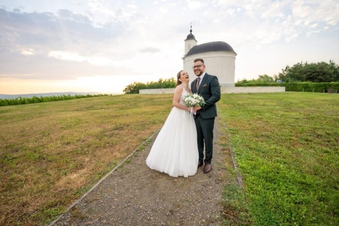 Egy vidám menyasszony és vőlegény áll a szabadban, egy kápolna előtt, háttérben a lenyugvó nap által megvilágított égbolttal. A menyasszony hófehér ruhában, virágcsokorral nevet, míg a vőlegény elegáns sötét öltönyben, barna nyakkendővel mosolyog mellette. A jelenet hangulatát a természet és a kápolna idilli látványa teszi különlegessé.