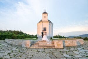 A menyasszony és a vőlegény állnak a Szent Teréz kápolna lépcsőinél, elegánsan felöltözve, miközben egymásra mosolyognak. A kápolna fehér homlokzata a háttérben kiemeli a jelenet romantikus és tiszta hangulatát. Az ég halvány színei harmonikusan illeszkednek a jelenethez, míg a menyasszony fátyla gyönyörűen szétterül a lépcsőkön.