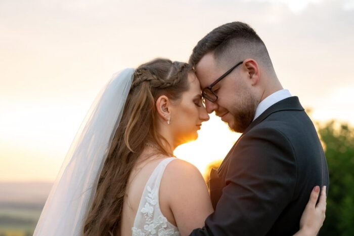 A menyasszony és a vőlegény meghitten állnak egymással szemben, homlokuk összeér. A naplemente lágy fénye megvilágítja arcukat, melegséggel és romantikával töltve meg a képet. A menyasszony hosszú fátyla finoman keretezi a kompozíciót, míg a vőlegény elegáns öltözékében a nyugalom és szeretet sugárzik.