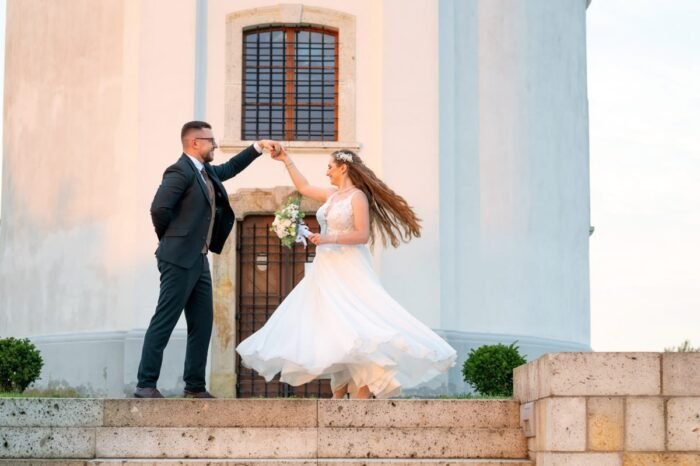 A menyasszony és a vőlegény elegáns táncmozdulatot mutatnak be a templom előtt, miközben a menyasszony ruhája kecsesen lebeg. A kép romantikus hangulatot áraszt, a háttérben pedig a templom részletei emelik ki a pillanat varázsát.