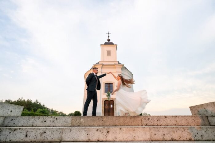 A menyasszony és a vőlegény kecses táncmozdulatot mutatnak be a templom lépcsőjén, miközben a menyasszony ruhája és haja a szélben lebeg. A háttérben a templom fehér épülete és a tiszta égbolt emeli ki a romantikus pillanatot.