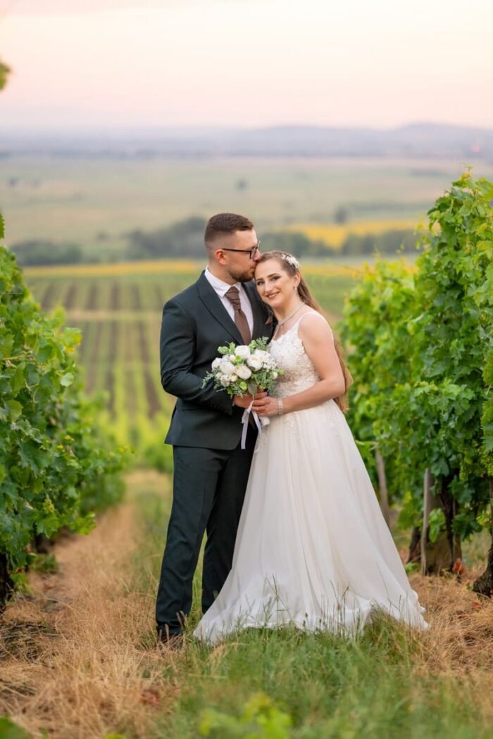 A menyasszony és a vőlegény szorosan egymáshoz bújva állnak egy szőlősor között, miközben a vőlegény gyengéden puszit ad a menyasszony fejére. A menyasszony mosolyogva tartja csokráját, a háttérben lágy naplemente és dombos táj látható.