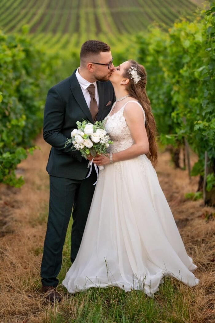 A menyasszony és a vőlegény szenvedélyes csókot vált egy szőlősorokkal ölelt mezőn. A menyasszony kezében fehér rózsákból és zöld növényekből álló csokor, a háttérben rendezett szőlőtőkék és a dombok láthatók.