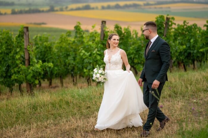 A menyasszony és a vőlegény mosolyogva sétálnak egy szőlősorokkal szegélyezett domboldalon. A menyasszony elegáns, csipkés menyasszonyi ruhát visel, kezében fehér rózsákból és zöld növényekből álló csokrot tart. A háttérben színes mezők és távoli hegyek nyújtanak festői látványt.