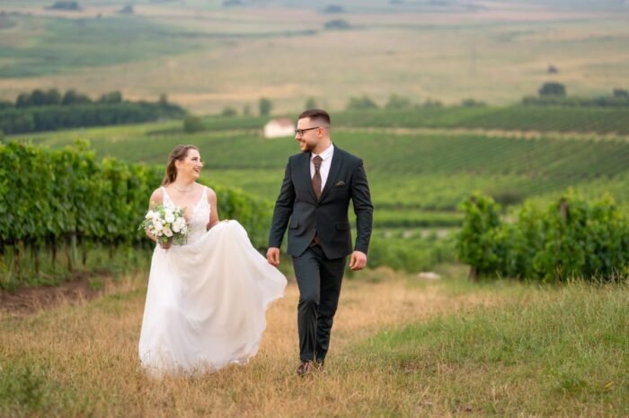 A boldog menyasszony és vőlegény mosolyogva sétálnak egymás mellett egy szőlőskertben. A menyasszony hófehér ruháját finoman megemelve tartja, kezében virágcsokrot visz, míg a vőlegény elegáns öltönyben követi. A háttérben a zöld szőlősorok és a távoli dombok látványa teszi idillivé a jelenetet.