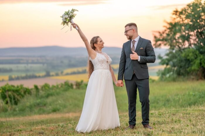 A boldog menyasszony egy kézzel magasba emeli a csokrot, miközben a vőlegény kezét fogja. Mindketten mosolyognak, mögöttük a naplemente festi aranyszínűre a tájat, a háttérben pedig dombok és mezők nyújtanak idilli hátteret a jelenethez.