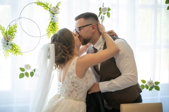 A menyasszony és a vőlegény gyengéd csókot váltanak egy romantikus dekorációval díszített helyiségben, ahol a természet ihlette virágdíszek és fényes ablakok teszik varázslatossá az esküvői pillanatot.