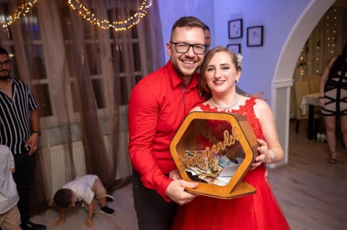 Az ifjú pár boldogan mosolyog, miközben kezükben tartják a nászajándékot, amelyet a vendégek adtak a menyecske tánc során. Az ünnepi hangulatot a háttérben lévő fényfüzérek és vidám vendégek teszik teljessé.