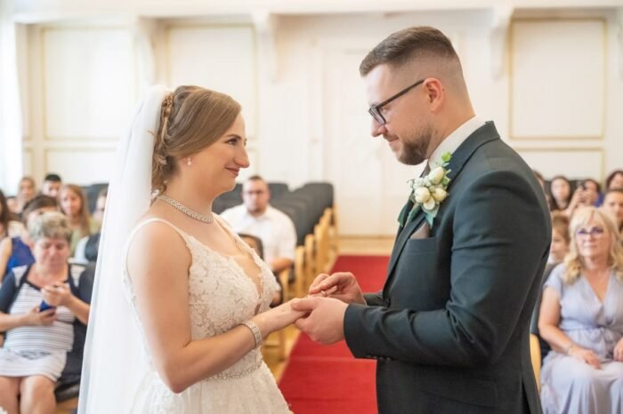 Az ifjú pár meghitt pillanatban, mosolyogva helyezik egymás ujjára a gyűrűt az esküvői szertartás során. A háttérben meghatódott vendégek figyelik az eseményt, miközben a terem elegáns hangulata emeli a pillanat ünnepélyességét.