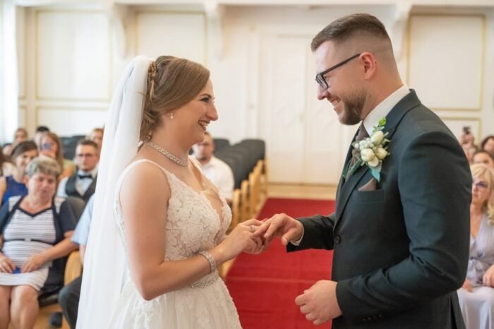 Az ifjú pár boldogan mosolyogva cseréli ki a gyűrűket az esküvői szertartás meghitt pillanatában. A menyasszony és a vőlegény között megjelenő szeretet és öröm megható látványt nyújt. A háttérben a meghatott vendégek figyelik az eseményt.