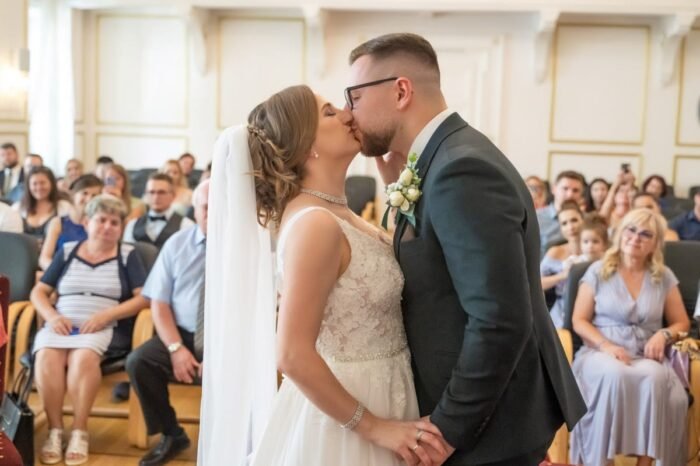 Az ifjú házaspár megosztja első csókját az esküvői szertartás után, miközben a háttérben vendégek mosolyognak és meghatódva figyelik a jelenetet. A menyasszony elegáns csipkeruhában, a vőlegény stílusos öltönyben áll.