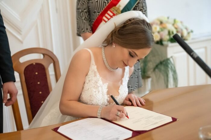 A menyasszony aláírja az esküvői hivatalos dokumentumot egy elegáns teremben. Fehér csipkeruhája és fátyla szépen keretezi az elmélyült pillanatot, míg a háttérben esküvői dekoráció és egy nemzeti szalag látható.