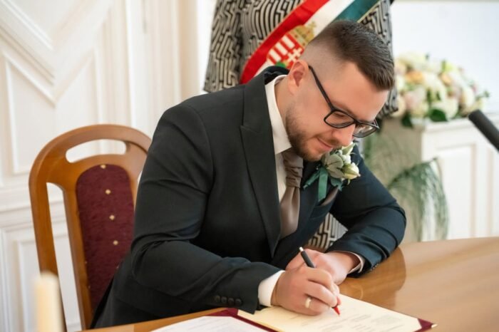 A vőlegény elegáns öltönyben, fehér ingben és nyakkendőben aláírja az esküvő hivatalos dokumentumát egy díszes teremben. Az asztalon esküvői papírok, a háttérben pedig virágdíszítés és nemzeti szalag látható.