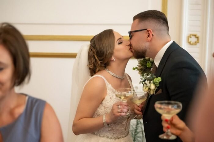 Az ifjú pár pezsgőspohárral a kezében csókolózik az esküvői ceremóniát követően. A menyasszony gyönyörű csipkeruhában, fátyollal, a vőlegény elegáns öltönyben és virágos kitűzővel látható. A háttérben és az előtérben esküvői vendégek tűnnek fel.