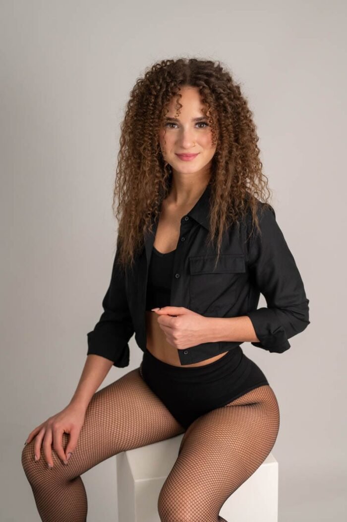 A nő fekete inget visel, miközben egy fehér kocka asztalon ül, fekete harisnyát és fekete bakancsokat viselve, mosollyal.