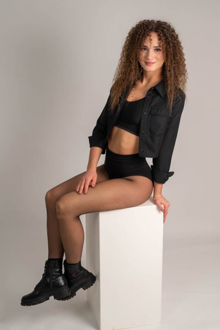 A nő fekete inget és harisnyát visel, miközben egy fehér kocka asztalon ül a stúdióban, fekete bakancsokkal és mosollyal.