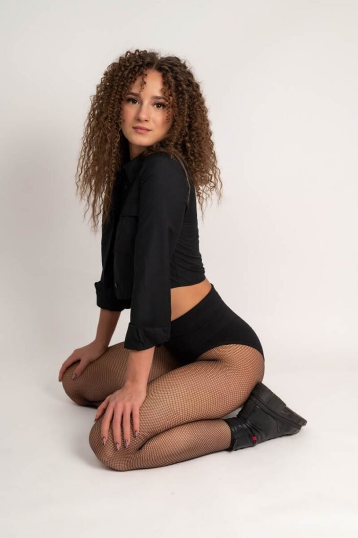 A nő fekete inget és harisnyát viselve térdel a stúdióban, miközben mosolyogva pózol fekete bakancsokban.