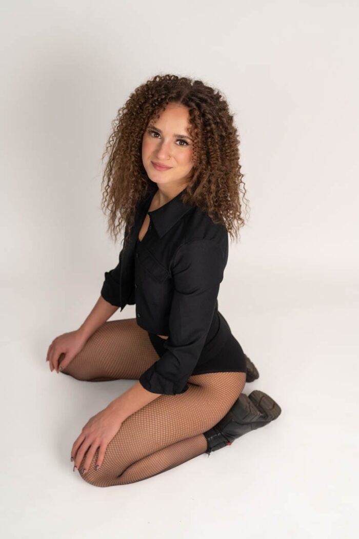 A nő fekete inget és harisnyát viselve térdel, mosolygós arccal és elegáns pózban, miközben a stúdióban ül.