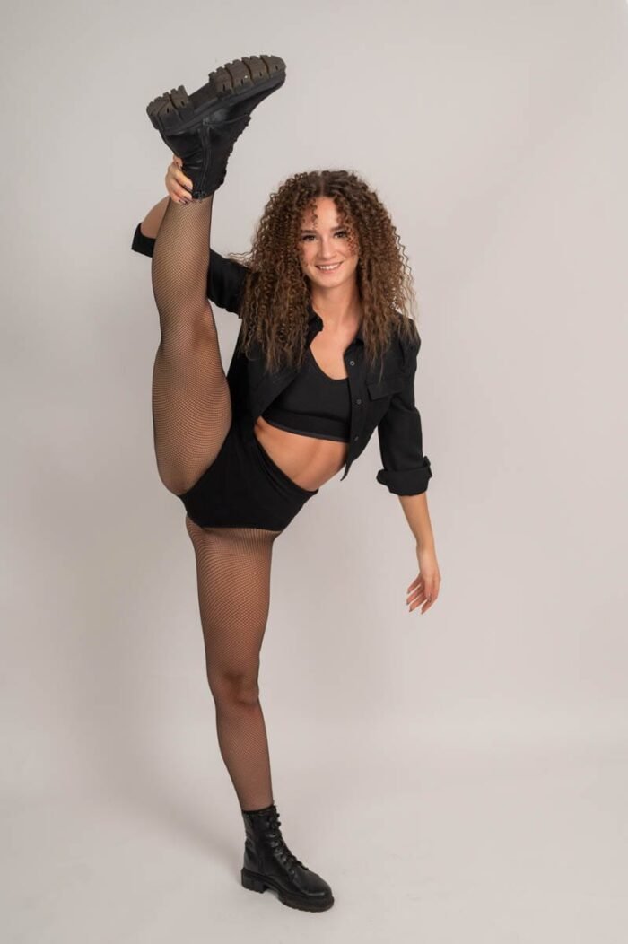 A fiatal nő fekete ingben, fekete harisnyában és bakancsban magasra emeli a lábát, miközben mosolyogva pózol a stúdióban