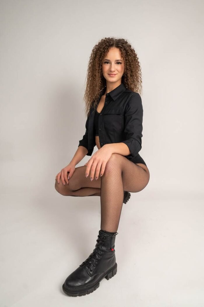 Fiatal nő fekete inget visel, fekete hálós harisnyát és fekete bakancsot, kicsit guggolva pózol a stúdióban.