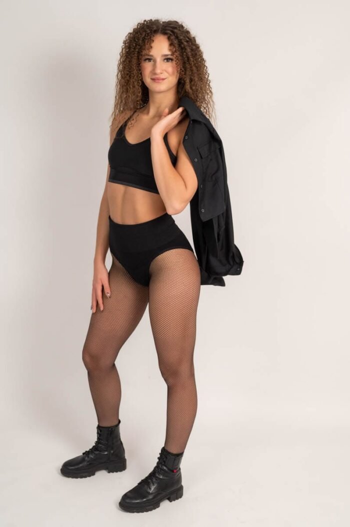 Fiatal nő fekete sportmelltartóban és magas derekú fekete nadrágban pózol, fekete inget tartva a vállán, stúdió környezetben. Fekete bakancs és hálós harisnya egészíti ki az outfitjét.