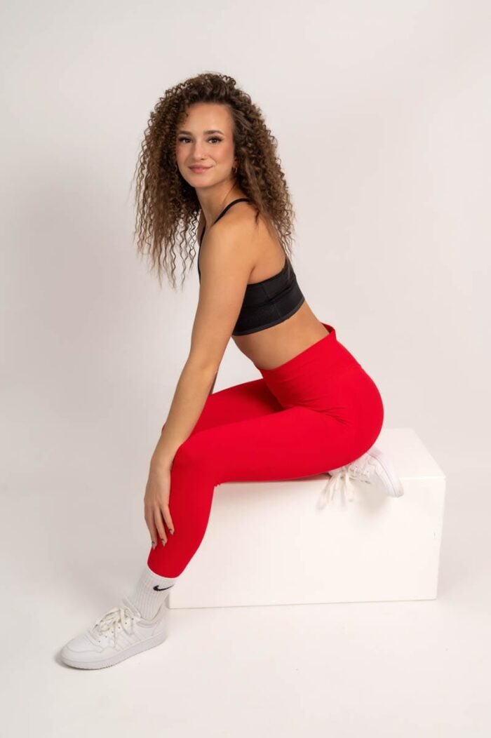 Nő piros sportnadrágban és fekete sportmelltartóban, fehér sportcipővel, fehér dobozon ülve, mosolygósan pózolva.