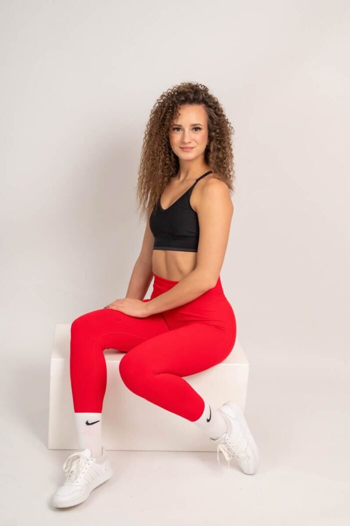 Nő piros sportnadrágban és fekete sportmelltartóban, fehér sportcipővel, egy fehér dobozon ülve, mosolygósan pózol.