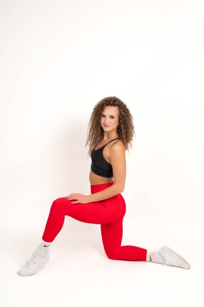 Nő piros sportnadrágban és fekete sportmelltartóban, fehér sportcipővel, térdelve, mosolygósan pózol.