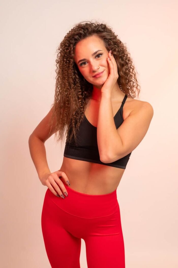 Nő piros sportnadrágban és fekete sportmelltartóban, fehér sportcipővel, egyik kezét az arcához emelve mosolyog a kamerába.