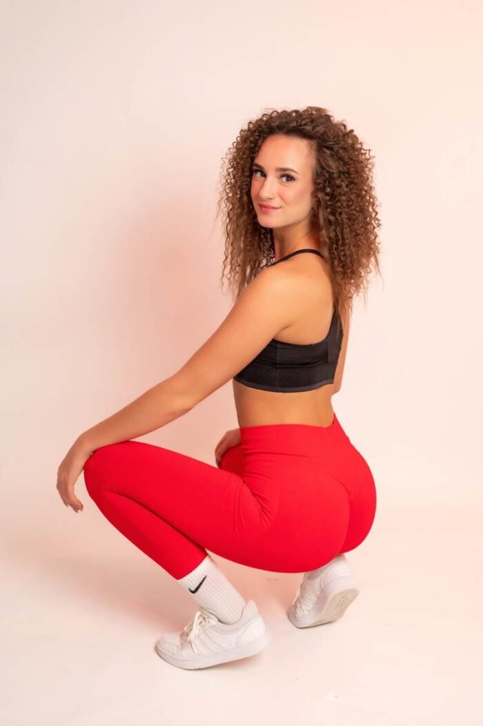 Nő piros sportnadrágban és fekete sportmelltartóban, fehér sportcipőben, guggoló pozícióban, oldalt nézve mosolyog a kamerába.