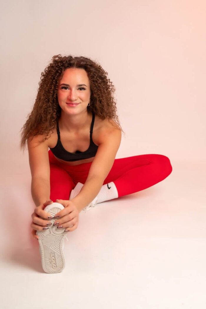 Nő piros sportnadrágban és fekete topban, fehér sportcipőben nyújtja a lábát, miközben mosolyogva néz a kamerába. Az egyik lábát kinyújtva, a cipőjét tartja.
