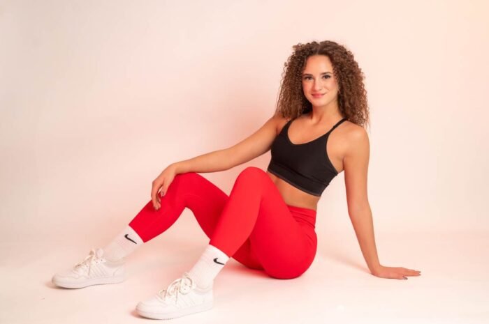 Fiatal nő piros sportnadrágban, fekete topban és fehér sportcipőben ül a földön. Mosolyogva néz a kamerába, egyik kezével a térdén, másikkal hátrafelé támaszkodik.