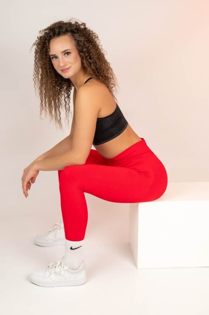 A modell egy piros sportnadrágban és fekete sportmelltartóban ül, miközben fehér sportcipőt és zoknit visel. Mosolyogva néz a kamerába, miközben egy fehér dobozra támaszkodik.