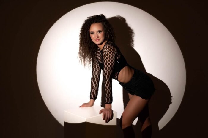 A képen egy nő látható, aki fekete hálós felsőt és rövidnadrágot visel. A modell egy fehér dobozon támaszkodik, miközben szemben áll a kamerával. A háttérben egy nagy kör alakú fényjáték található, amely kiemeli a modell alakját és arcát. A nő fekete bakancsot visel, és mosolyogva néz a kamerába.