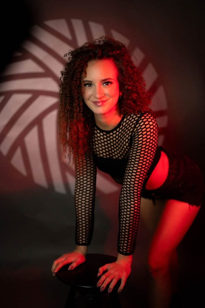 A képen egy nő látható, aki fekete hálós felsőt és rövidnadrágot visel. Kezeit egy szék háttámláján pihenteti, miközben szemben áll a kamerával. A háttérben egy geometrikus fényjáték látható, piros fényekkel, amelyek egy érdekes árnyékot vetnek.