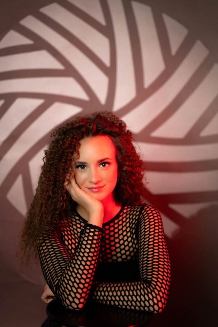 A képen egy nő látható, aki fekete hálós felsőt visel, kezét az arcán pihenteti, míg egy piros fényekkel megvilágított geometrikus mintázatú háttér előtt ül. A nő arckifejezése nyugodt és mosolygós, haját gondosan formázva, a fények pedig kontrasztot adnak a portrénak, kiemelve a textúrát és az alakját.