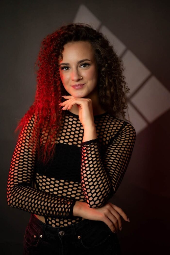 A képen egy fiatal nő látható, aki egy fekete hálós felsőt visel, amely kiemeli a modern, egyedi stílust. A háttér sötét, és piros fényekkel világítva, drámai hatást kelt. A nő magabiztosan mosolyog, miközben egyik kezét az állán tartja, a másik kezét a csípőjén. A fények és a kompozíció erőteljes vizuális hatást biztosítanak, és kiemelik a nő arcát és öltözetét.