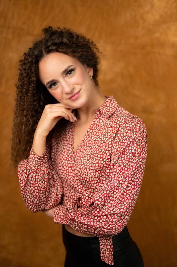 Modell fotózás Debrecenben. A képen egy fiatal nő látható, aki piros-fehér virágmintás blúzt visel. A háttér narancssárga, meleg tónusú, amely kiemeli a nő természetes szépségét. A nő egy kezet a fejére támaszt, miközben mosolyogva néz a kamerába. A finom fények és a lágy háttér biztosítják, hogy a fókusz a nő arcán maradjon, a mosolygós és magabiztos arckifejezés egyedi hangulatot ad a fotónak.