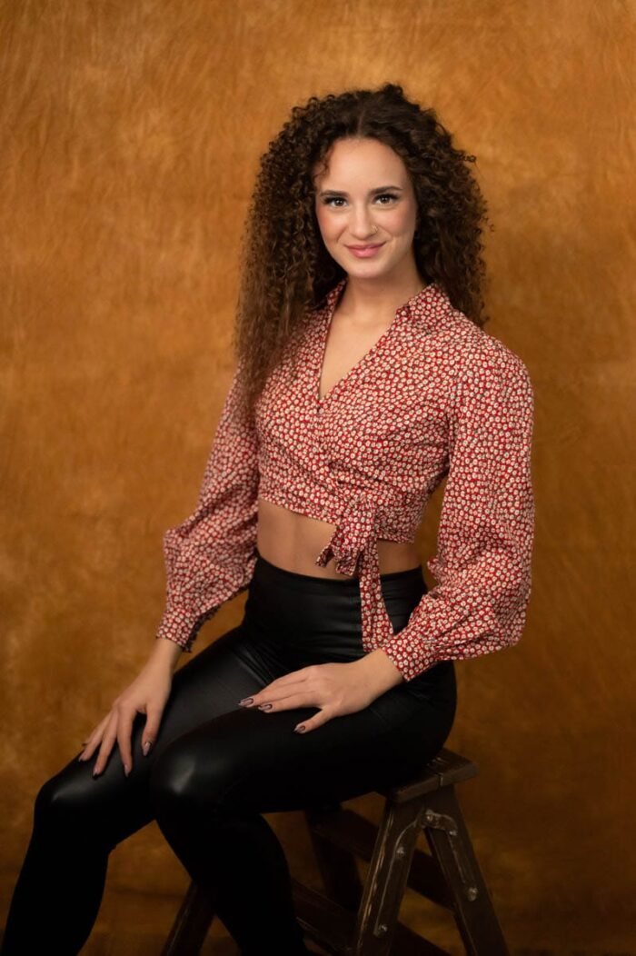 A képen egy fiatal nő látható, aki egy létrán ül. Fekete, bőrszerű nadrágot és fekete bakancsot visel, miközben piros-fehér mintás blúzát a derekánál kötötte meg. A háttér narancssárga színű, amely meleg hangulatot ad a fotónak. A nő mosolyogva néz a kamerába, természetes és barátságos arckifejezéssel. Az arcát finom fények világítják meg, kiemelve a vonásait, miközben kényelmesen és magabiztosan pózol.