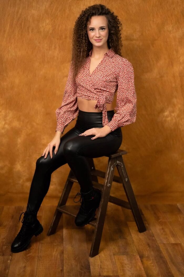 A képen egy fiatal nő látható, aki egy létrán ül, fekete bőrnadrágot és fekete bakancsot visel, miközben egy piros-fehér mintás blúzt visel, amely kiemeli a derekát. A háttér narancssárga színű, ami kellemes, meleg hangulatot ad a fotónak. A nő mosolyogva néz a kamerába, kezét nyugodtan pihenve tartja a térdén, miközben barátságos és természetes megjelenést mutat. A fények finoman megvilágítják az arcát, kiemelve a vonásait.